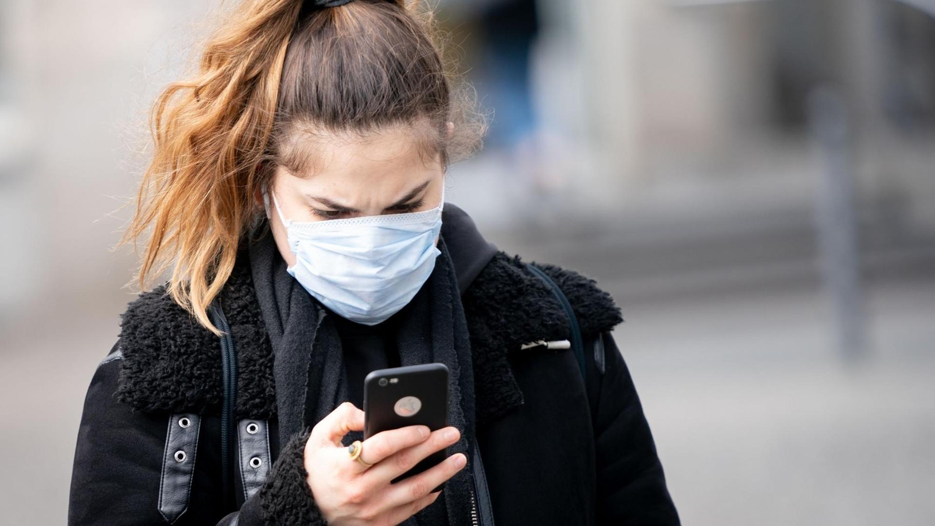 Eine Frau mit Mund-Nasen-Maske blickt auf ihr Smartphone. Eine Frau mit Mund-Nasen-Maske blickt auf ihr Smartphone.