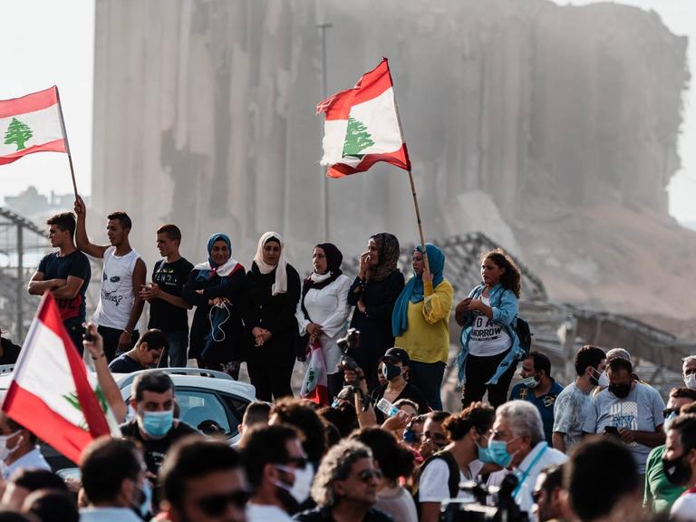 Menschen haben sich vor dem zerstörten Hafen versammelt, sie schwenken Nationalflaggen
