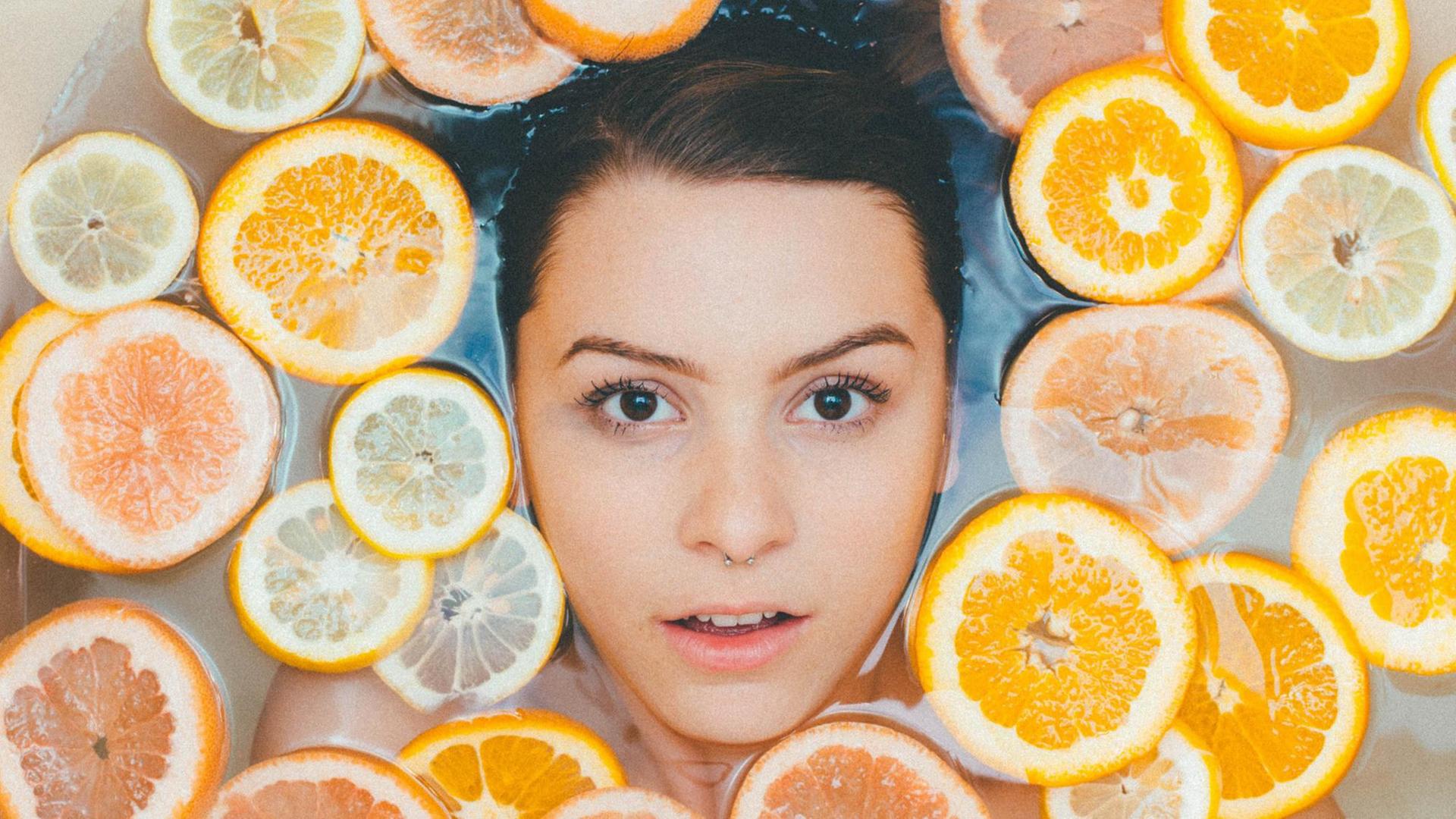 Der Kopf einer Frau mit Nasenpiercing von oben in der Badewanne liegend fotografiert, um sie herum schwimmen Orangen- und Zitronenscheiben