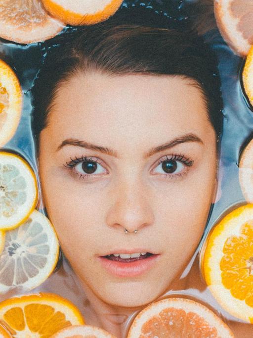 Der Kopf einer Frau mit Nasenpiercing von oben in der Badewanne liegend fotografiert, um sie herum schwimmen Orangen- und Zitronenscheiben