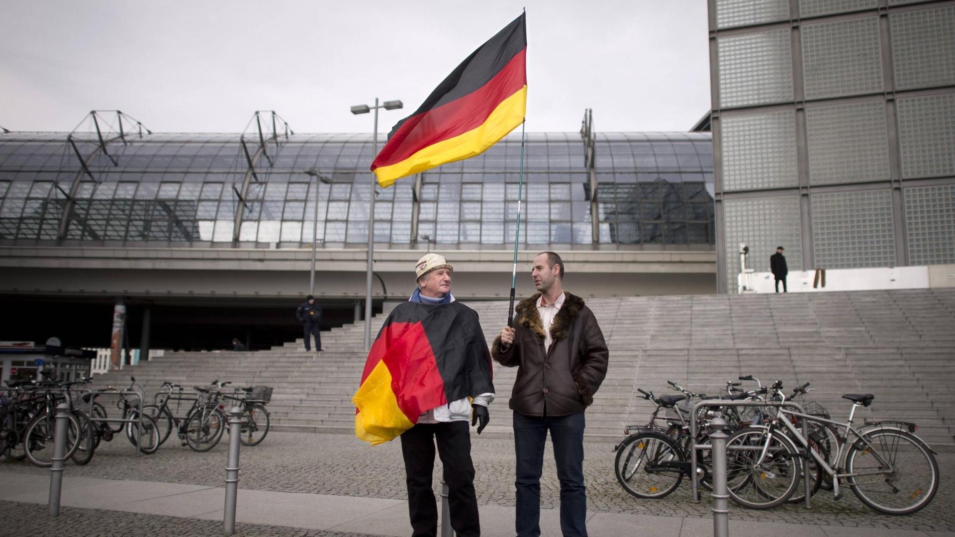 Zwei Männer stehen vor einer grauen Steintreppe, einer hält eine Deutschland-Fahne in der Hand, der andere hat eine Deutschland-Fahne um den Oberkörper gewickelt. Zwei Männer stehen vor einer grauen Steintreppe, einer hält eine Deutschland-Fahne in der Hand, der andere hat eine Deutschland-Fahne um den Oberkörper gewickelt.
