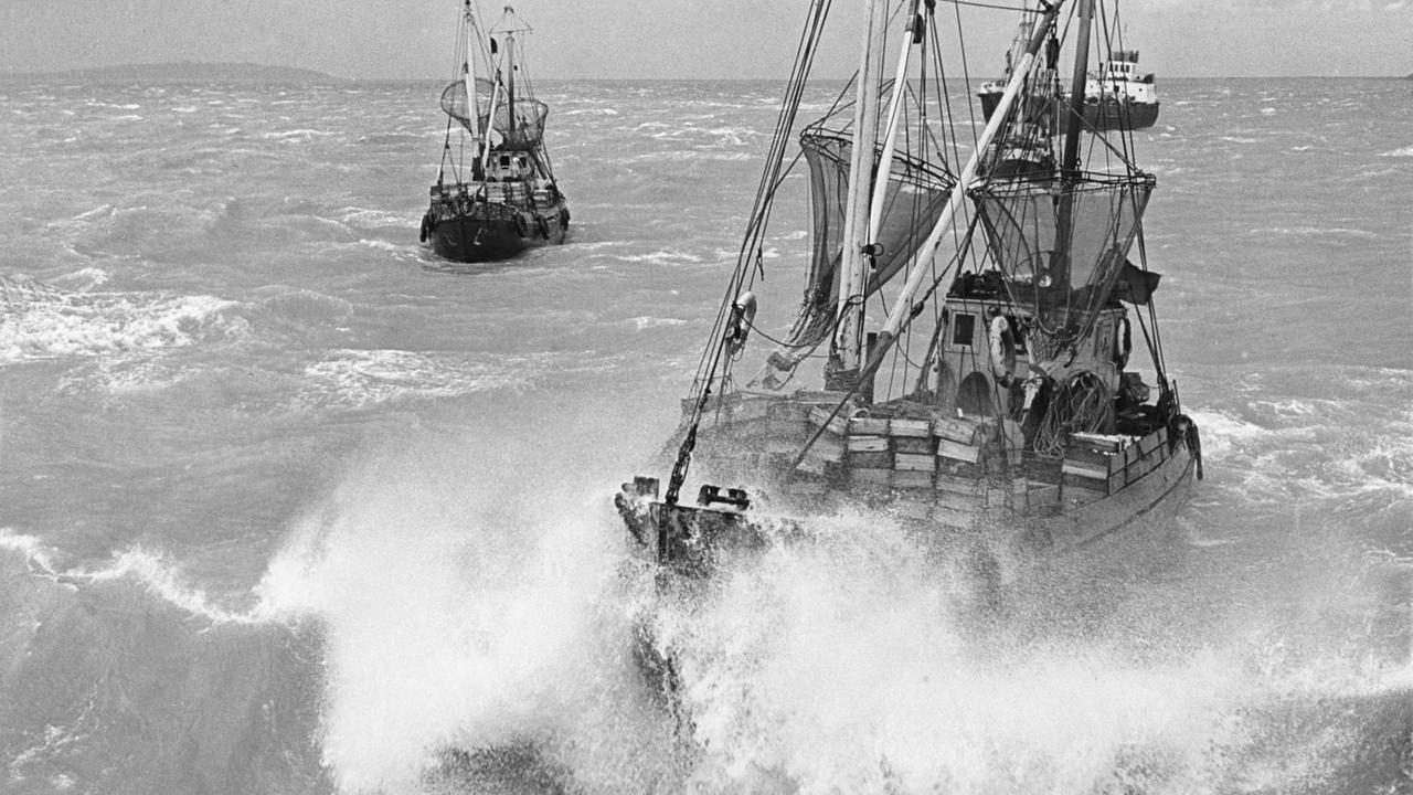 Zwei große Fischerboote mit hohen Masten und eingerollten Segeln befinden sich inmitten tosender Wellen. Zwei große Fischerboote mit hohen Masten und eingerollten Segeln befinden sich inmitten tosender Wellen.