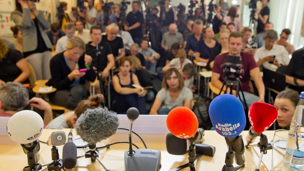 Sie sehen bunte Mikrofone und im Hintergrund viele Journalisten. Sie sehen bunte Mikrofone und im Hintergrund viele Journalisten.