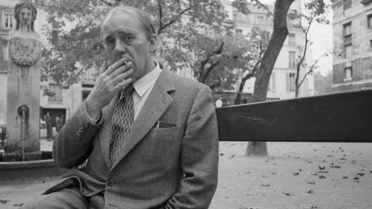 Heinrich Böll sitzt auf einer Bank, raucht und schaut dabei direkt in die Kamera.