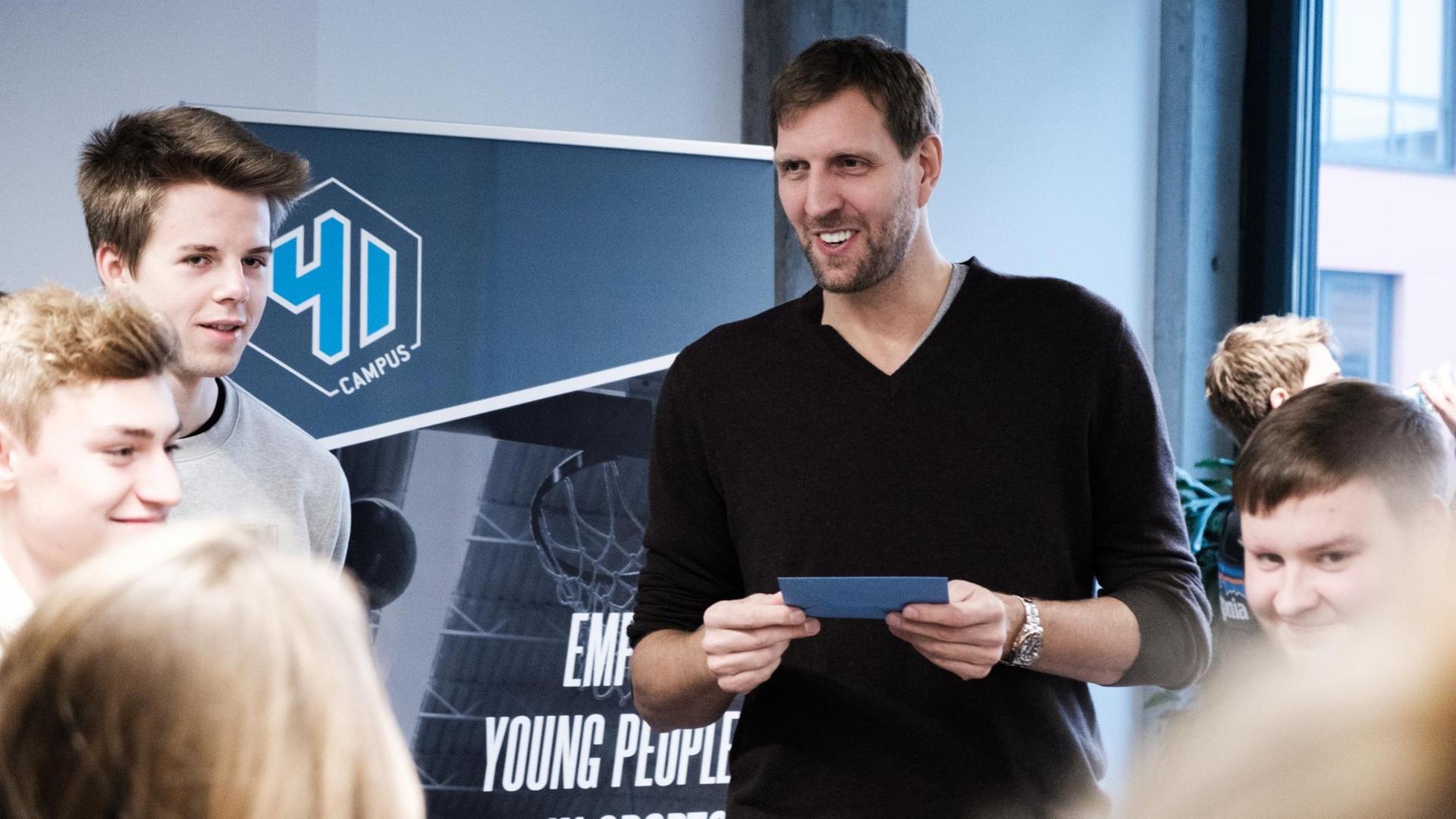 Jugendliche stehen im Halbkreis um Dirk Nowitzki, die eine blaue Moderationskarte in seinen Händen hält. Jugendliche stehen im Halbkreis um Dirk Nowitzki, die eine blaue Moderationskarte in seinen Händen hält.