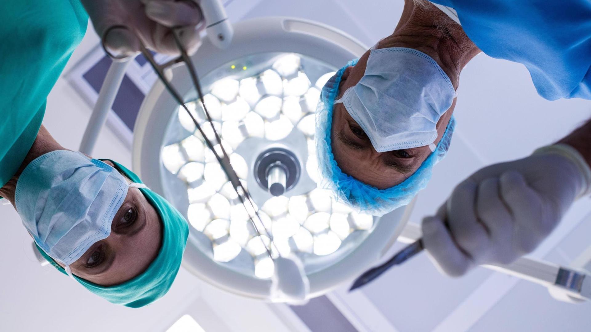 Zwei Ärzte beugen sich unter typischen Op-Strahlern über den möglichen Patienten. Zwei Ärzte beugen sich unter typischen Op-Strahlern über den möglichen Patienten.