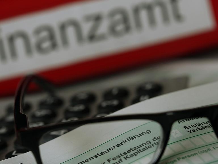 Auf einem Vordruck für die Einkommensteuererklärung liegen vor einem Aktenordner mit der Aufschrift "Finanzamt" eine Brille und ein Kugelschreiber