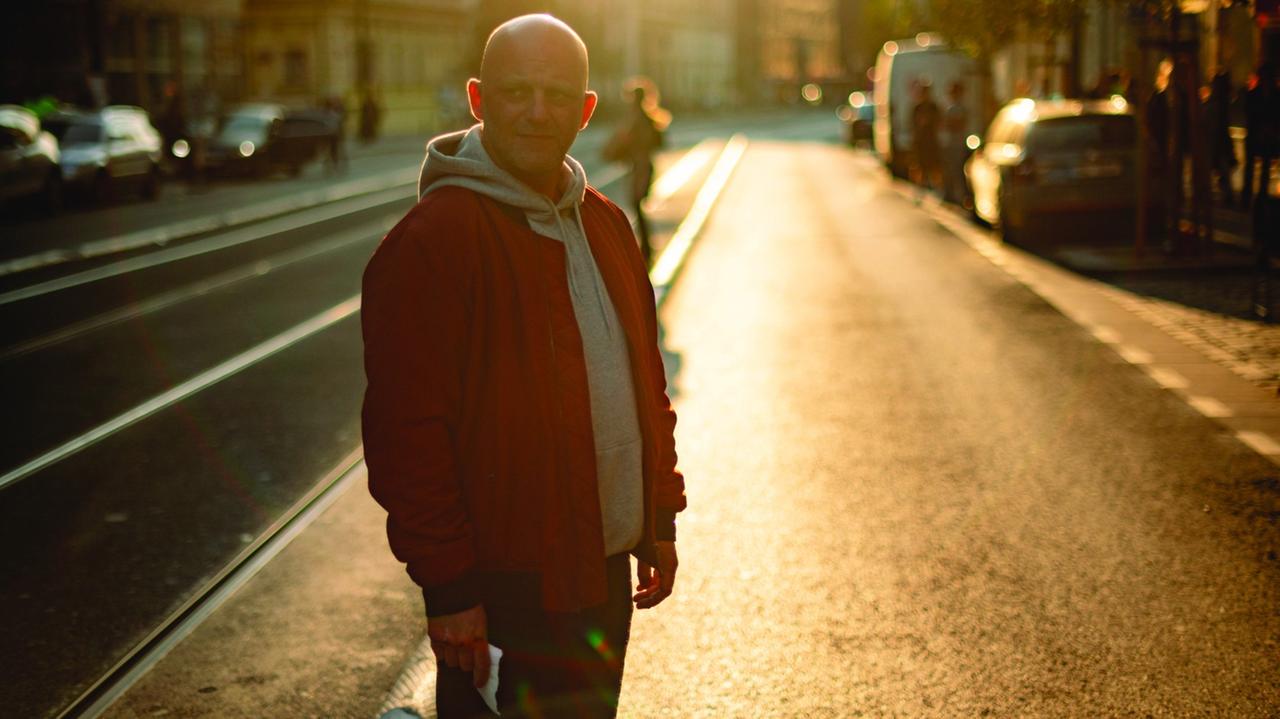 Ein Mann in Skinheadmontur steht im Gegenlicht einer Abend- oder Morgensonne auf einer Straße.
