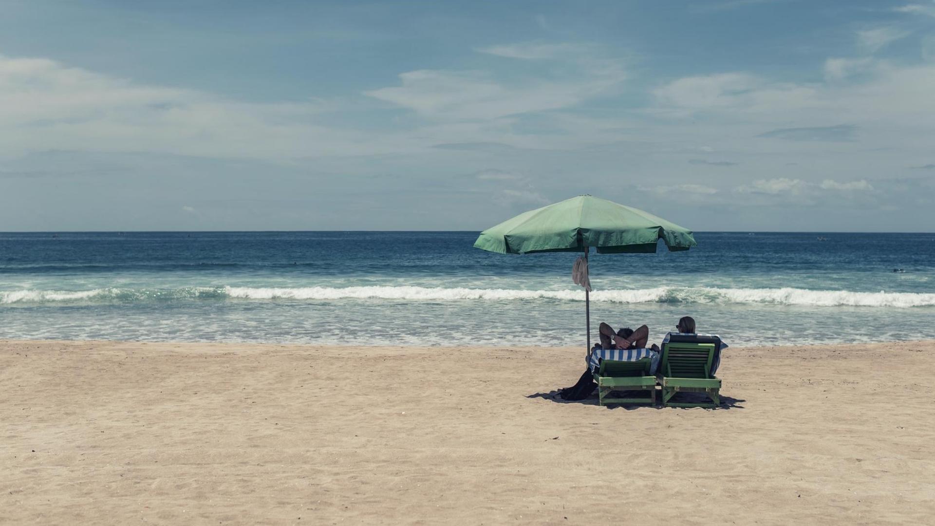 Bild eines fast komplett leeren Strands mit Blick aufs Meer: Nur zwei Menschen liegen auf ihren Strandliegen unter einem Sonnenschirm. Bild eines fast komplett leeren Strands mit Blick aufs Meer: Nur zwei Menschen liegen auf ihren Strandliegen unter einem Sonnenschirm.