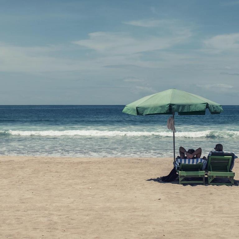 Bild eines fast komplett leeren Strands mit Blick aufs Meer: Nur zwei Menschen liegen auf ihren Strandliegen unter einem Sonnenschirm. 