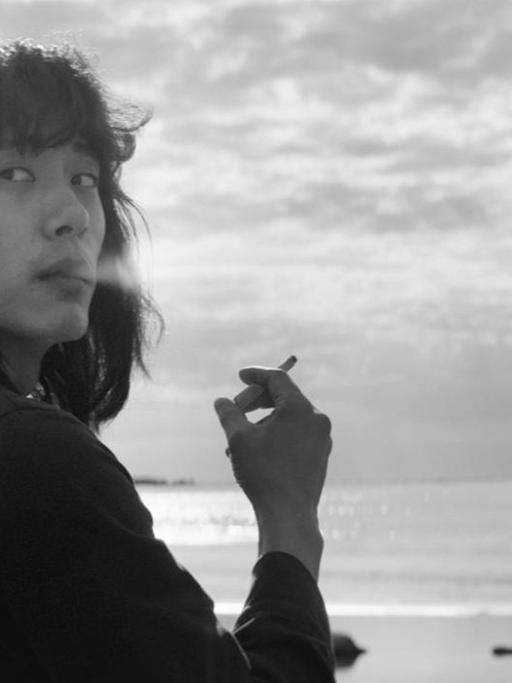 Schwarzaufnahme eines asiatischen Schauspielers, der Zigarette rauchend am Strand steht und nachdenklich über seine Schulter blickt.