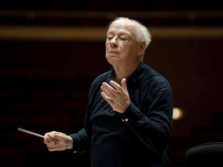 Der Dirigent Bernard Haitink steht auf einer dunklen Bühne und dirigiert ein nicht sichtbares Symphonie Orchester.