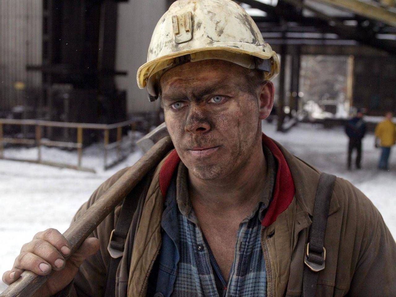 Das Bild zeigt einen Bergarbeiter, der mit einem verrußtem Gesicht und einem Hammer über der Schulter vor der Kamera steht.