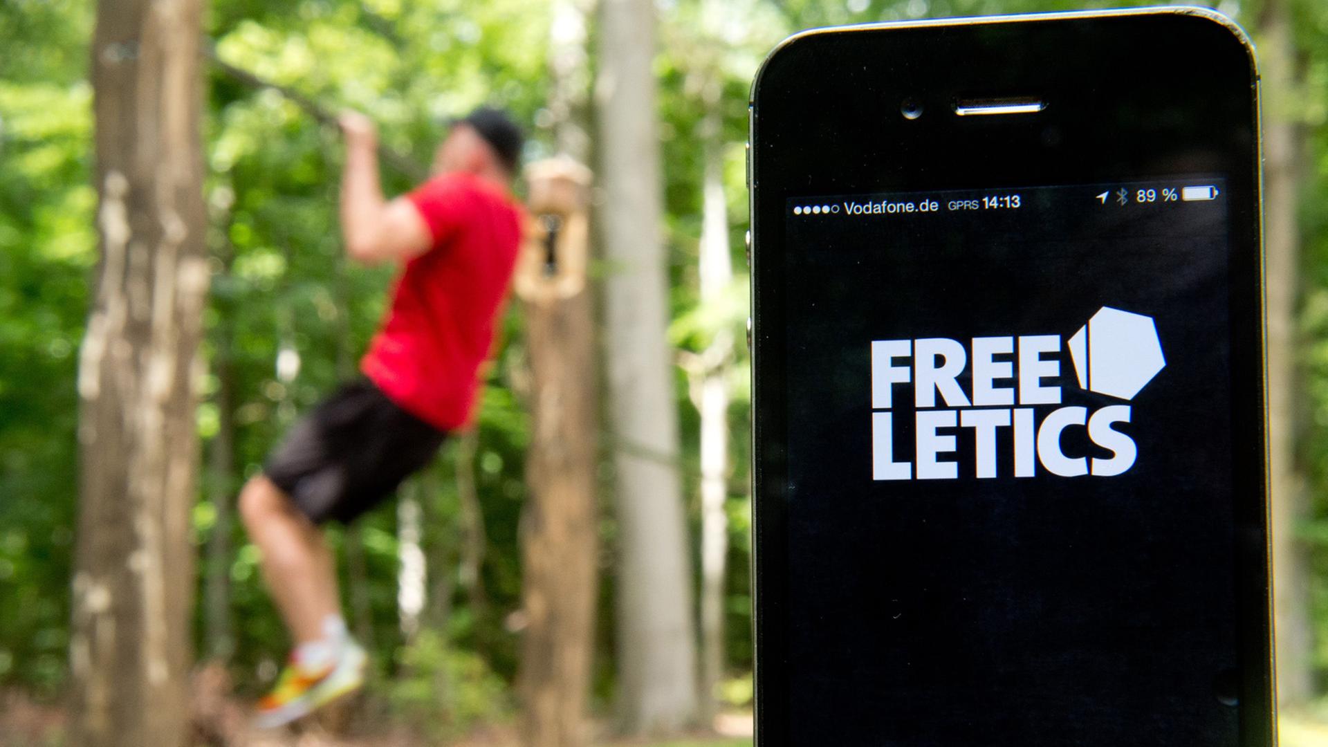 Die Fitness-App "Freeletics" wird auf einem iPhone angezeigt, während im Hintergrund ein Mann auf einem Trimm-Dich-Pfad einen Klimmzug macht. Die Fitness-App "Freeletics" wird auf einem iPhone angezeigt, während im Hintergrund ein Mann auf einem Trimm-Dich-Pfad einen Klimmzug macht.