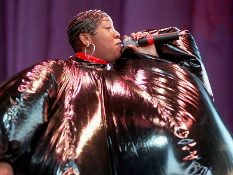 Eine gestylte schwarze Frau mit einem Mikrofon in der Hand. SIe trägt ein überdimensionales schwarz-pinkes Kostüm, das aussieht wie ein aufgeblasener Ballon.