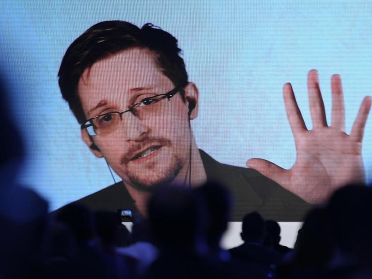 Über verschatteten Sitzreihen scheint ein auf die Bühne projizierter Stream auf, über den Edward Snowden an einer Konferenz teilnimmt.