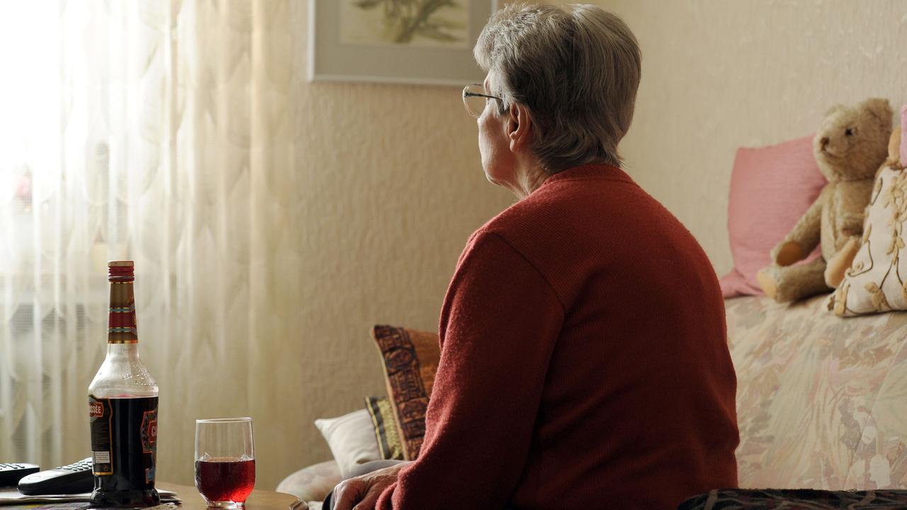 Eine alte Frau sitzt einsam in ihrer Wohnung vor einer Flasche Alkohol