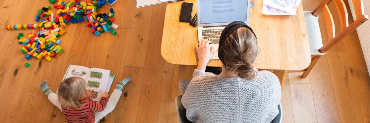Ein zweijähriges Kind spielt im Wohnzimmer, während seine Mutter Zuhause im Homeoffice an einem Laptop arbeitet. Ein zweijähriges Kind spielt im Wohnzimmer, während seine Mutter Zuhause im Homeoffice an einem Laptop arbeitet.
