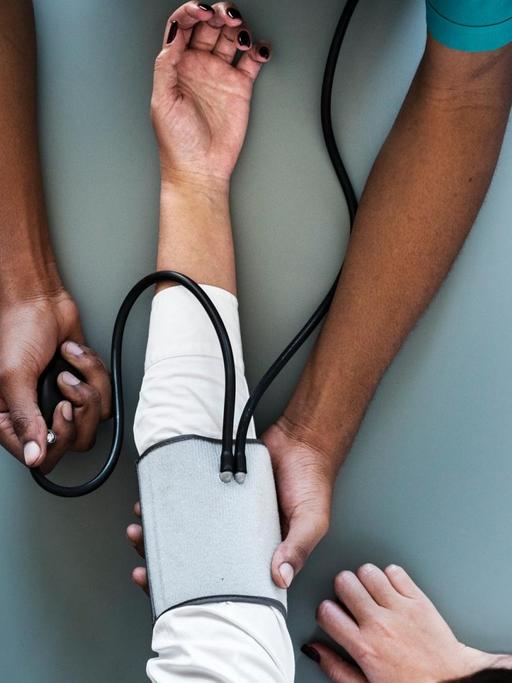 Eine Pflegekraft misst den Blutdruck eines Patienten. Von beiden sind nur die Hände und Arme zu sehen.