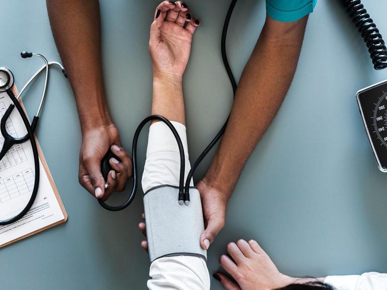 Eine Pflegekraft misst den Blutdruck eines Patienten. Von beiden sind nur die Hände und Arme zu sehen.