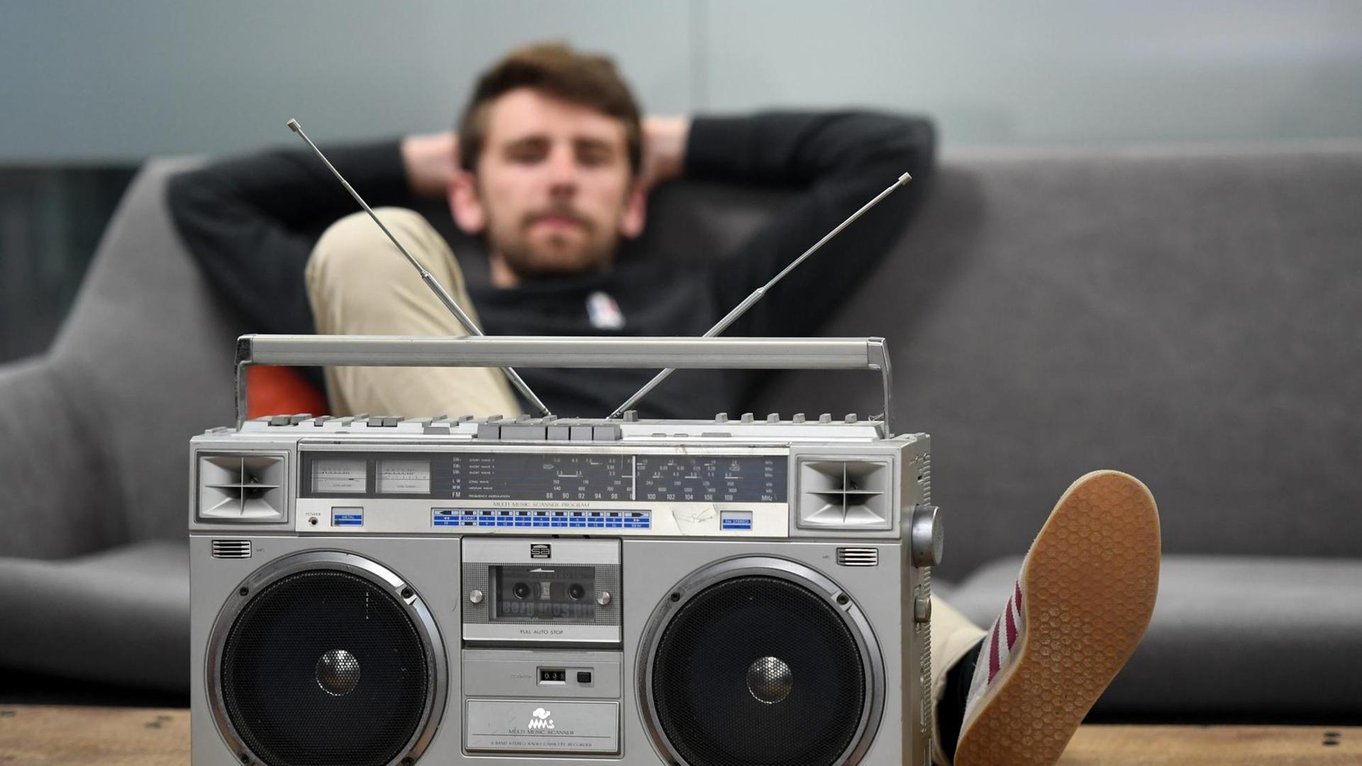 Ein dunkelhaariger Mann sitzt auf einem grauen Sofa und hat einen Fuß auf dem Couchtisch abgelegt. Vor ihm ein altmodischer Kassettenrekorder. Ein dunkelhaariger Mann sitzt auf einem grauen Sofa und hat einen Fuß auf dem Couchtisch abgelegt. Vor ihm ein altmodischer Kassettenrekorder.