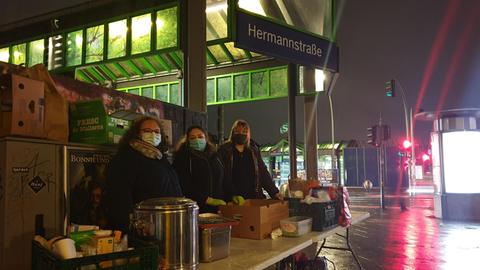 Unter einem S-Bahnschild mit der Aufschrift "Hermannstraße" stehen drei Frauen hinter einem Campingtisch, auf dem Essen aufgebaut ist.