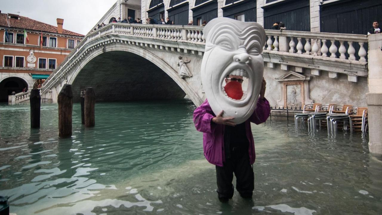 Ein Mann mit einer gigantischen Maske steht im Hochwasser nahe der Rialto-Brücke während der außergewöhnlichen Flut am 17. November 2019. Ein Mann mit einer gigantischen Maske steht im Hochwasser nahe der Rialto-Brücke während der außergewöhnlichen Flut am 17. November 2019.