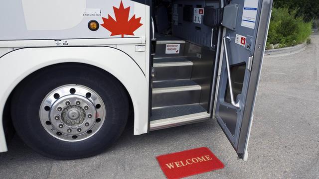 Ein Greyhound Bus mit einem roten Ahornblatt als Zeichen für Kanada. Die Fahrertür ist offen, darunter liegt eine Fußmatte auf der Erde, auf der "Welcome" zu lesen ist.