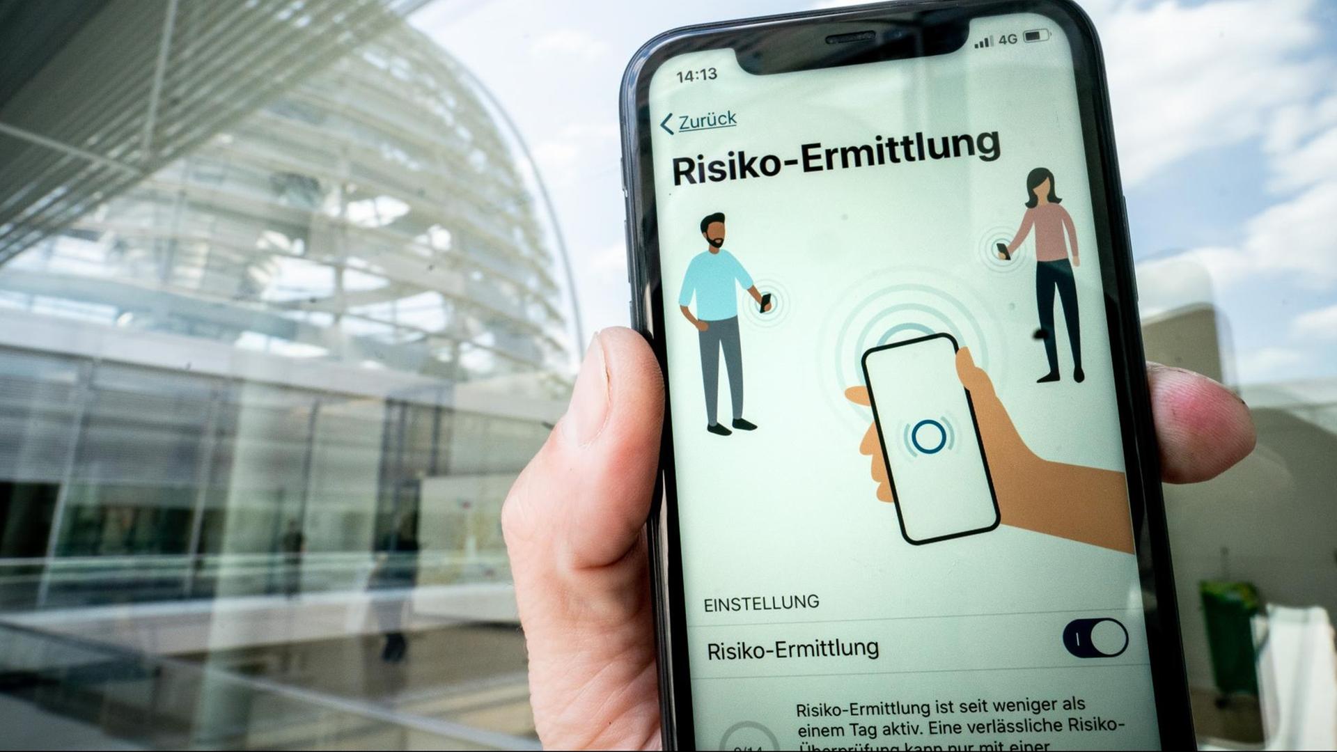 Die Corona-Warn-App mit der Seite zur Risiko-Ermittlung ist im Display eines Smartphone vor der Kuppel des Reichstags zu sehen.