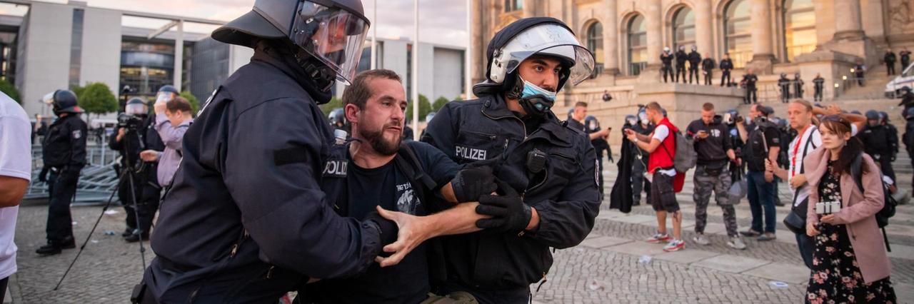 Zwei Polizisten führen den in schwarz gekleideten Demonstranten vor den Treppen zum Reichstag ab. Er scheint sich zu wehren. Dahinter weitere Polizisten und Demonstranten Zwei Polizisten führen den in schwarz gekleideten Demonstranten vor den Treppen zum Reichstag ab. Er scheint sich zu wehren. Dahinter weitere Polizisten und Demonstranten