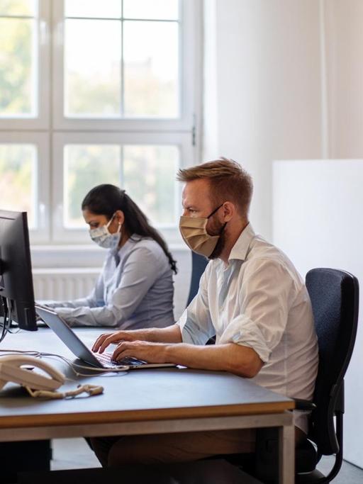 Zwei Arbeitnehmer mit Mundschutz, die während der Pandemie im Büro am Schreibtisch arbeiten.