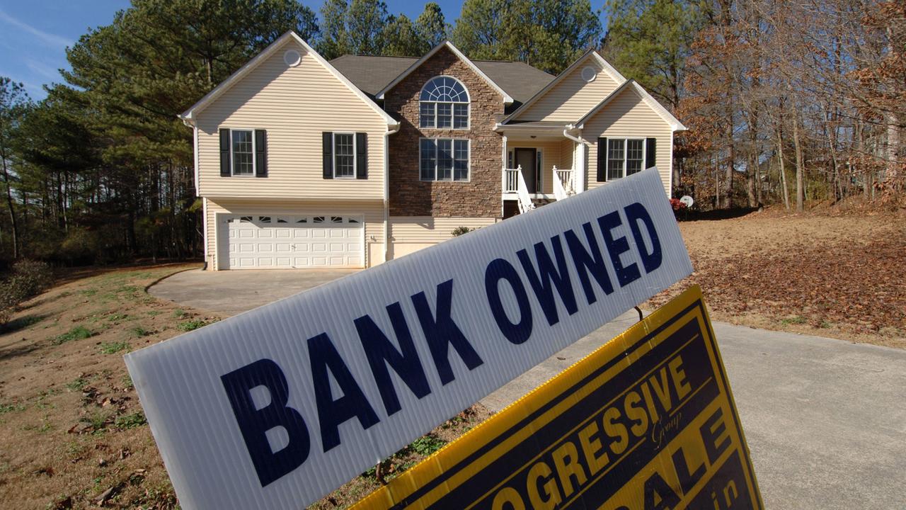Ein mit Holz verkleidetes Haus in den USA, im Vordergrund ein Schild, auf dem "Bank owned" steht.