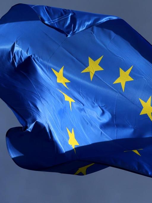 Die sonnenbeschienene blaue Flagge mit dem gelben Sternenkreis flattert vor einem dunklen Himmel.