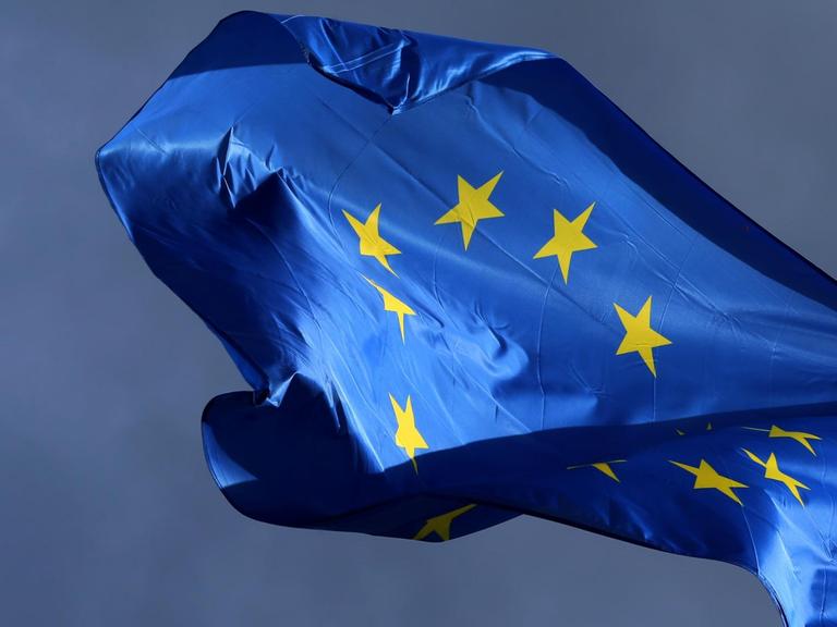 Die sonnenbeschienene blaue Flagge mit dem gelben Sternenkreis flattert vor einem dunklen Himmel.