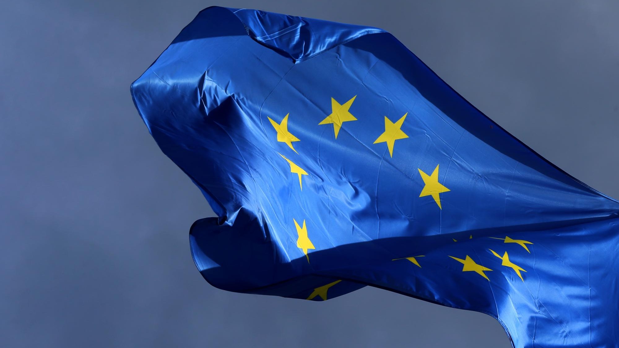 Die sonnenbeschienene blaue Flagge mit dem gelben Sternenkreis flattert vor einem dunklen Himmel.