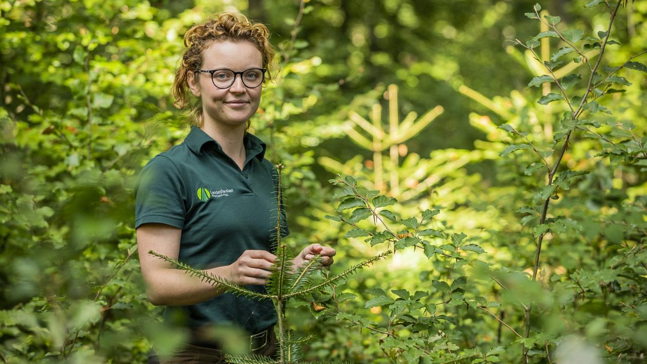Anne-Sophie Knop steht in grüner im Wald zwischen jungen Nadelbäumen Anne-Sophie Knop steht in grüner im Wald zwischen jungen Nadelbäumen