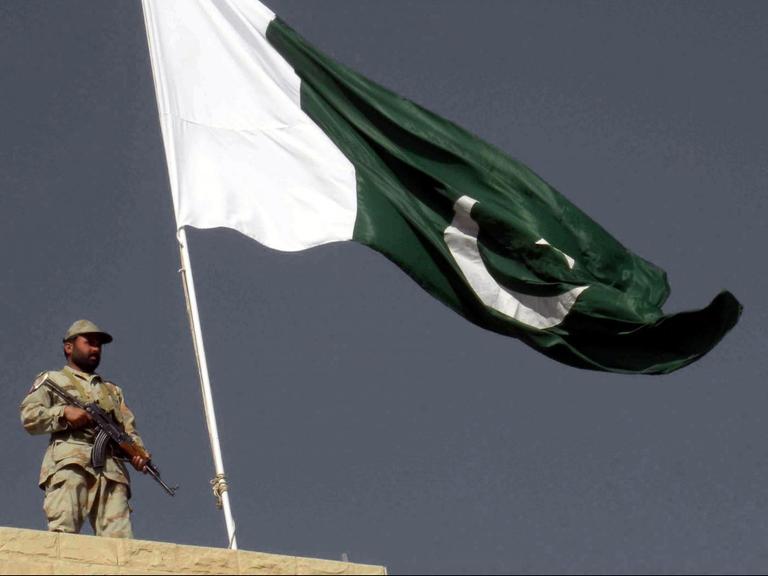 Ein Pakistanischer Soldat steht auf einer Mauer an der pakistanisch-afghanischen Grenze. Im Hintergrund weht die Pakistanische Flagge