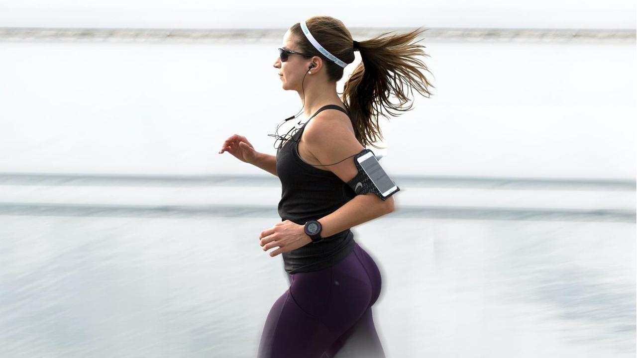 Eine Frau in Sportbekleidung joggt mit einem am Arm befestigten Smartphone. Eine Frau in Sportbekleidung joggt mit einem am Arm befestigten Smartphone.
