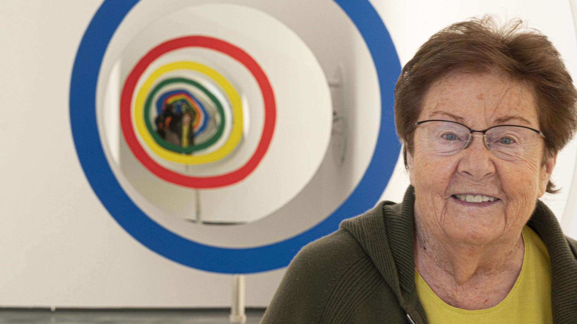 Eine ältere Frau mit kurzen Haaren steht in einem Museum vor einem Kunstwerk und lächelt in die Kamera. Eine ältere Frau mit kurzen Haaren steht in einem Museum vor einem Kunstwerk und lächelt in die Kamera.