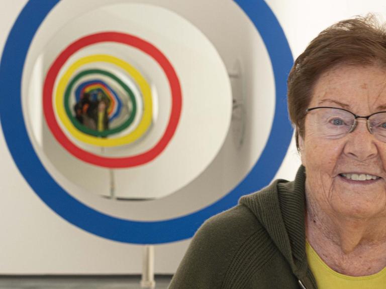 Eine ältere Frau mit kurzen Haaren steht in einem Museum vor einem Kunstwerk und lächelt in die Kamera.