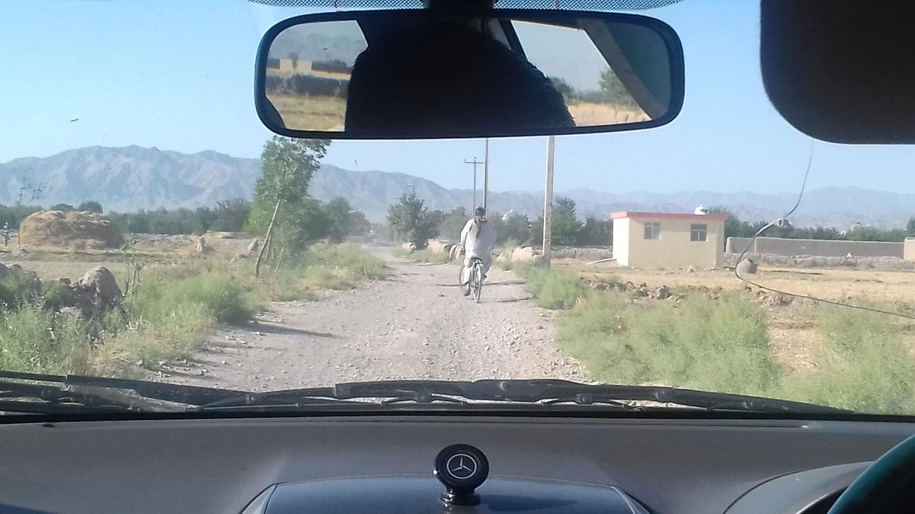 Auf dem Weg zum Safran-Garten: Blick aus dem fahrenden Auto auf eine staubige Schoterpiste, vor dem Wagen ein Fahrradfahrer im langen weißen Gewand. Auf dem Weg zum Safran-Garten: Blick aus dem fahrenden Auto auf eine staubige Schoterpiste, vor dem Wagen ein Fahrradfahrer im langen weißen Gewand.