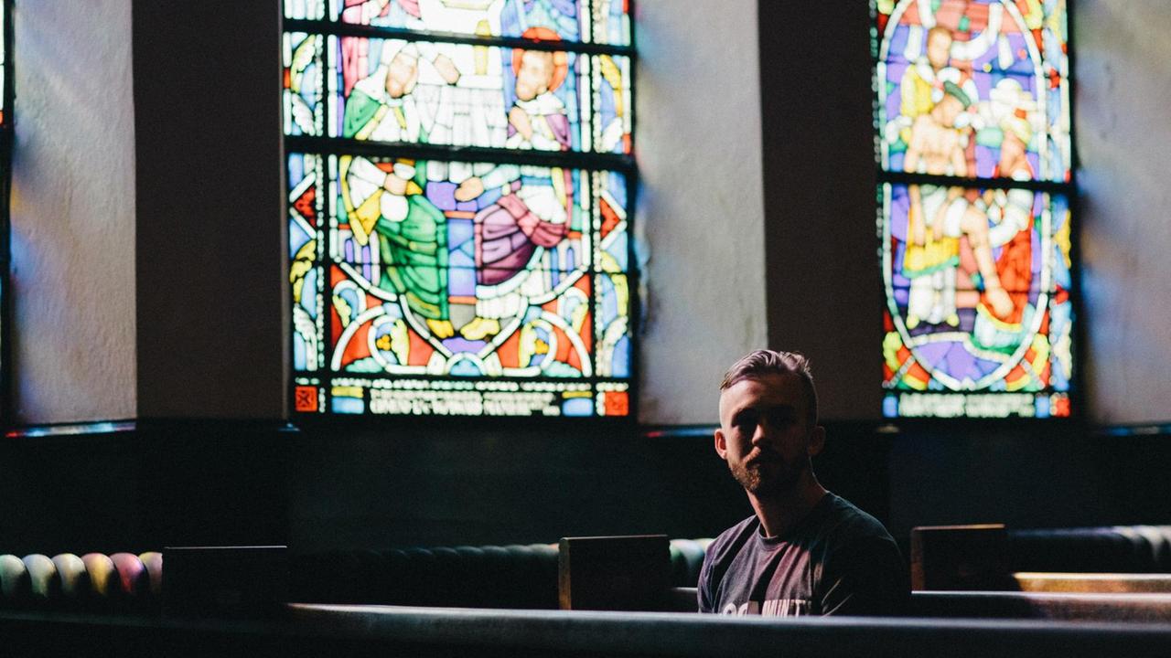 Ein junger Mann sitzt alleine auf einer Kirchenbank, im Hintergrund sind bunte Kirchenfester zu sehen. Ein junger Mann sitzt alleine auf einer Kirchenbank, im Hintergrund sind bunte Kirchenfester zu sehen.