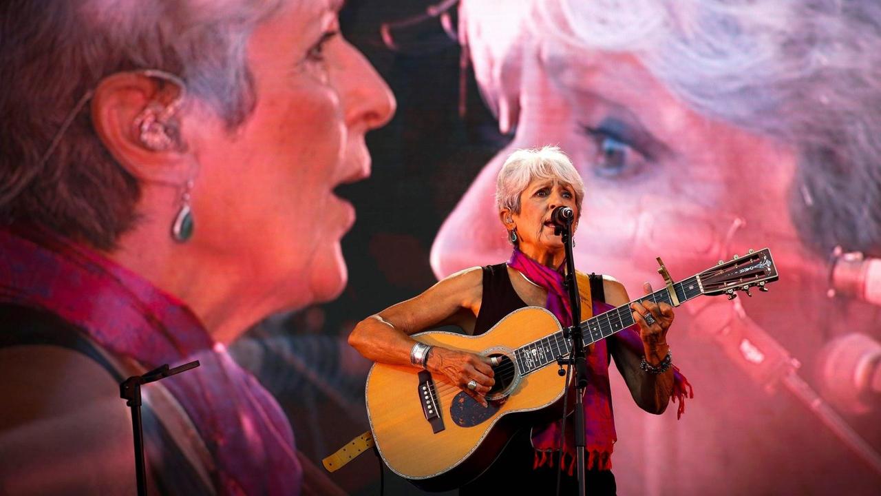 Eine Sängerin mit kurzem, grauen Haar steht mit ihrer Gitarre auf der Bühne, während ihr Porträt zwei Mal übergroß auf Leinwänden hinter ihr erscheint. Eine Sängerin mit kurzem, grauen Haar steht mit ihrer Gitarre auf der Bühne, während ihr Porträt zwei Mal übergroß auf Leinwänden hinter ihr erscheint.
