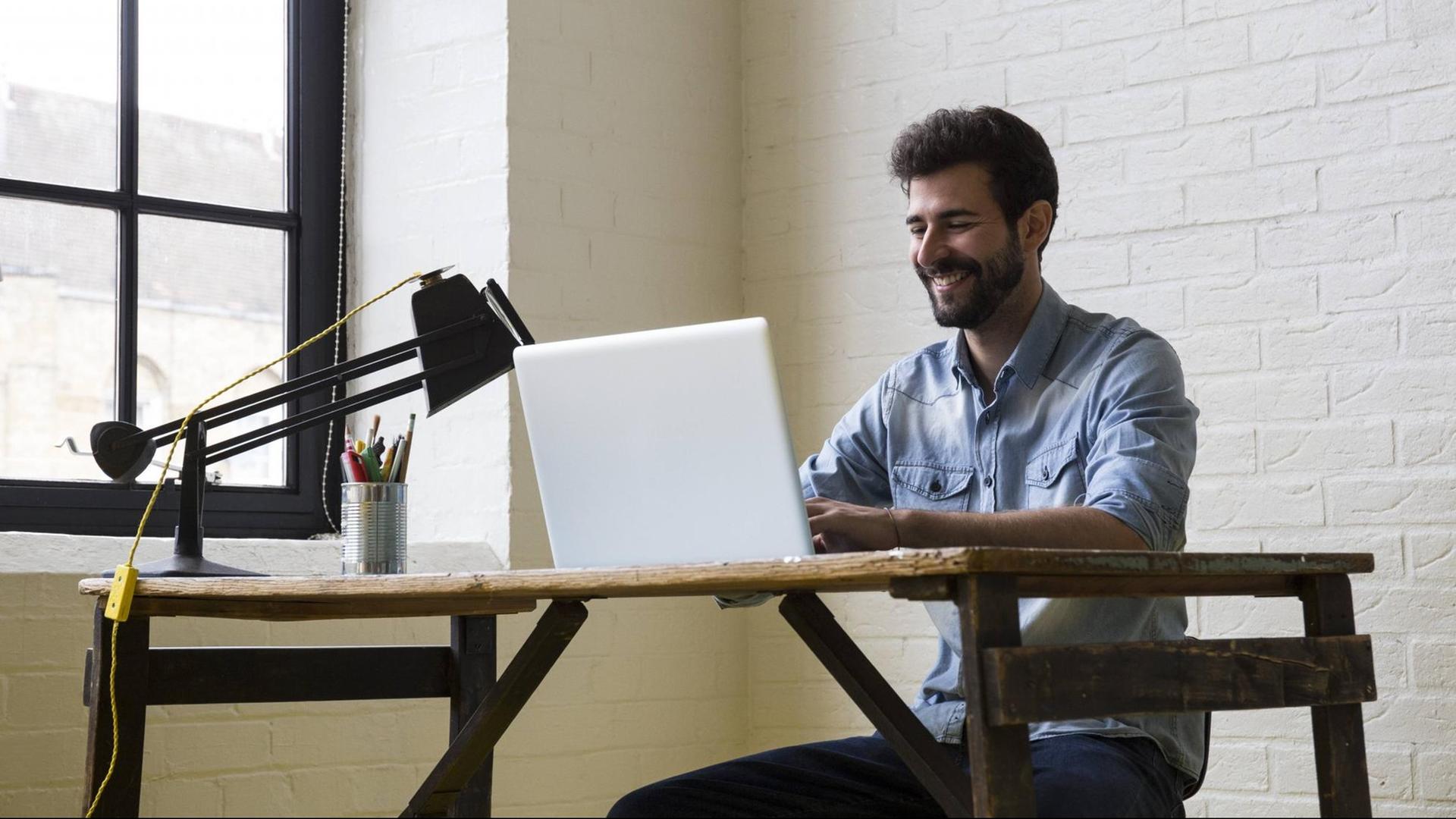 Ein lächelnder Mann sitzt an einem Schreibtisch, vor ihm steht ein Laptop Ein lächelnder Mann sitzt an einem Schreibtisch, vor ihm steht ein Laptop
