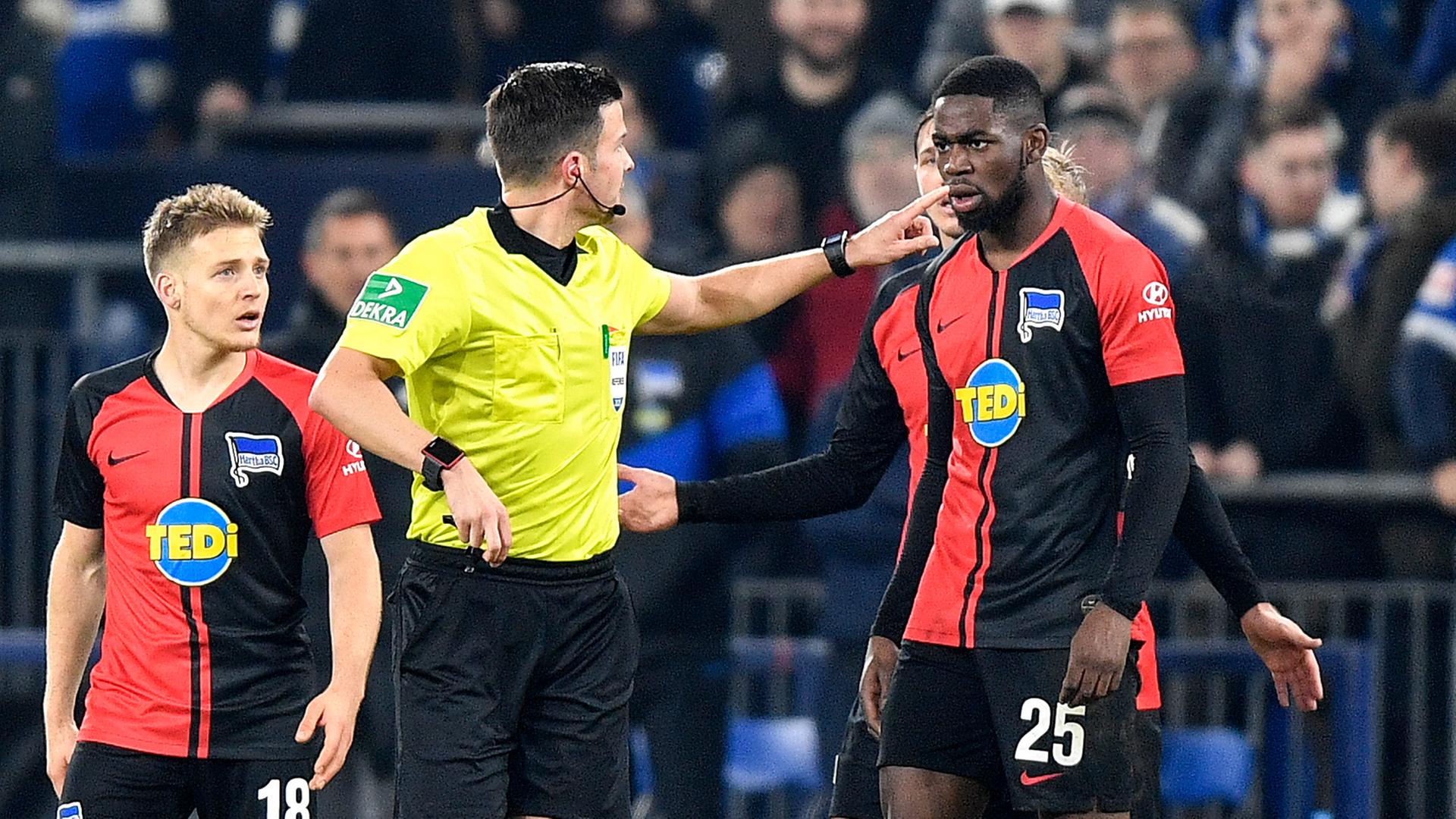 Osmers im gelben Schiedsrichter-Trikot zeigt mit dem Finger nach hinten, flankiert ist er von den beiden Hertha-Spielern im schwarz-roten Trikot. Osmers im gelben Schiedsrichter-Trikot zeigt mit dem Finger nach hinten, flankiert ist er von den beiden Hertha-Spielern im schwarz-roten Trikot.