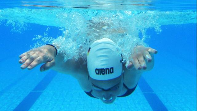 Eine Sportschwimmerin taucht mit weißer Badekappe ins lichtblaue Wasser.