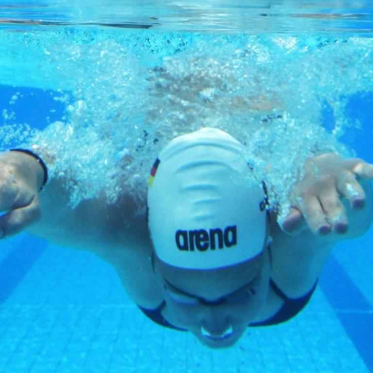 Eine Sportschwimmerin taucht mit weißer Badekappe ins lichtblaue Wasser.