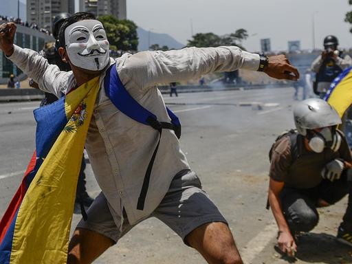 Ein Demonstrant, der eine Guy-Fawkes-Maske trägt und eine venezolanische Flagge um den Hals hat, wirft während einer Auseinandersetzung mit Sicherheitskräften Steine.