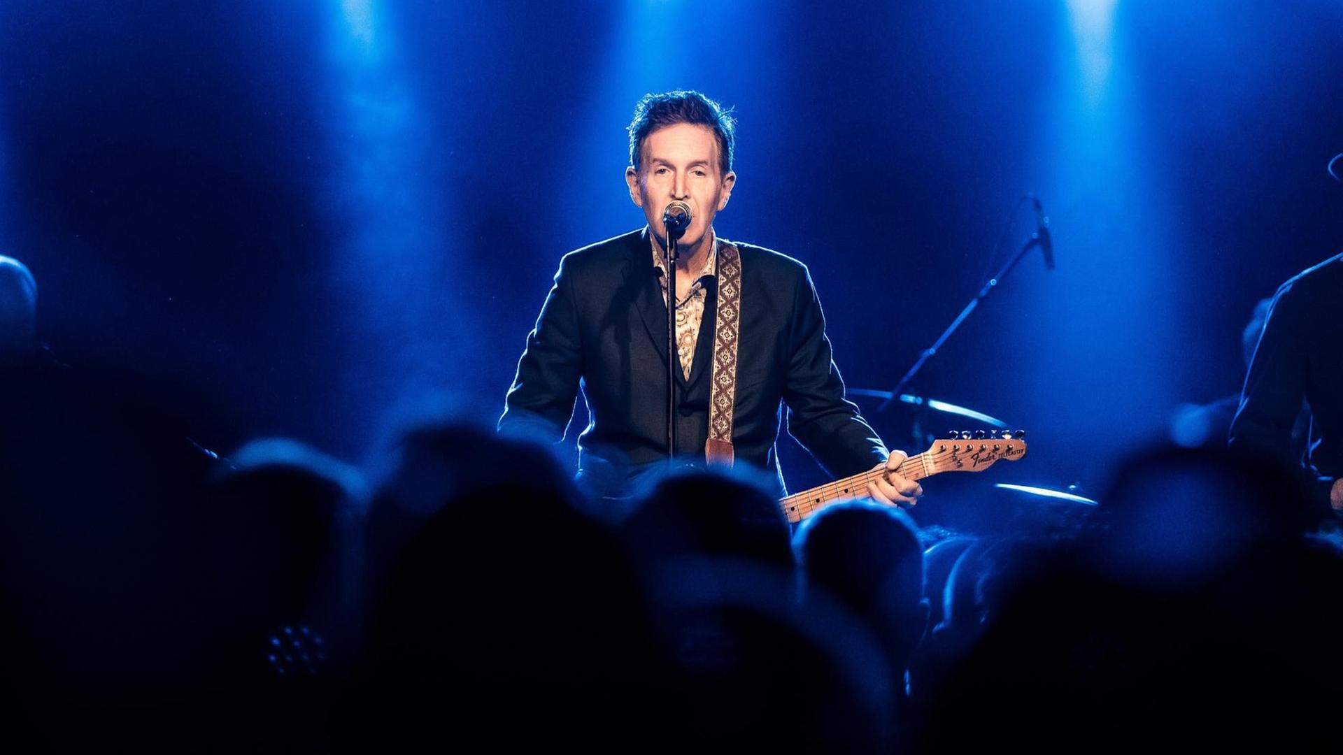 Der Sänger Steve Wynn auf der Bühne mit Gitarre und Mikrofon. Vor ihm in der Unschärfe das Publikum. Der Sänger Steve Wynn auf der Bühne mit Gitarre und Mikrofon. Vor ihm in der Unschärfe das Publikum.