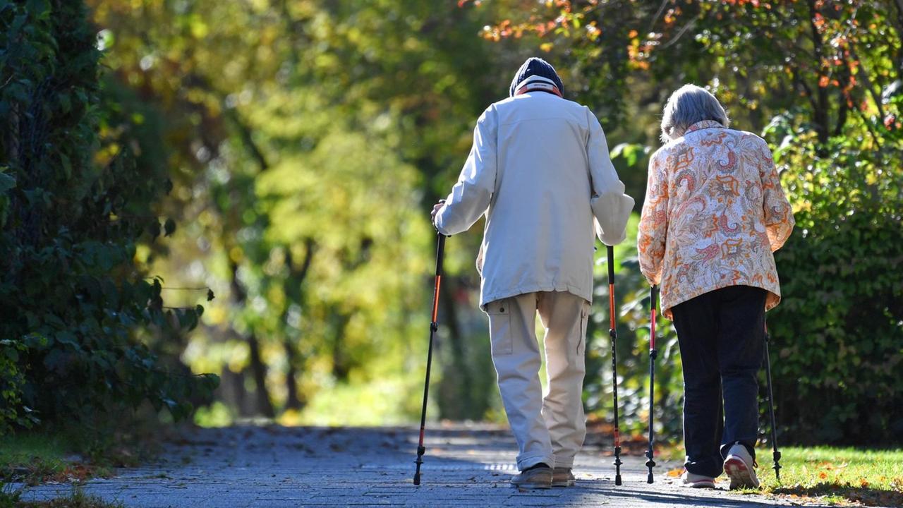 Zu sehen ist ein altes Rentner-Ehepaar, dass mit Nordic-Walking-Stöcken spazieren geht.